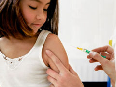 A preschool-aged girl in a tank top looks on as a health care provider gives her a shot in the upper arm