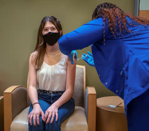 A teenage girl sits in a chair while a healthcare worker administers the coronavirus vaccine to her upper arm