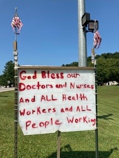 A large white sign on poles on grass in Jackson, Ky. with small American flags taped to both poles. The sign is written in red spray paint and says "God Bless our Doctors and Nurses and ALL Health Workers and ALL People Working